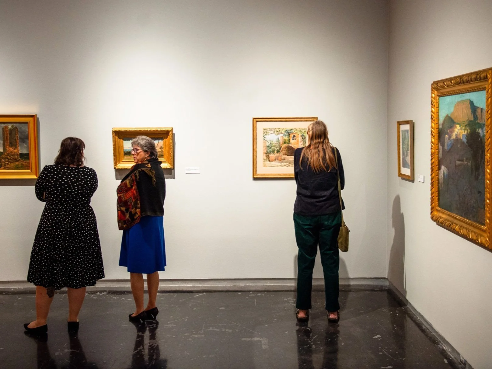 Visitors observing framed paintings on the walls of a gallery.