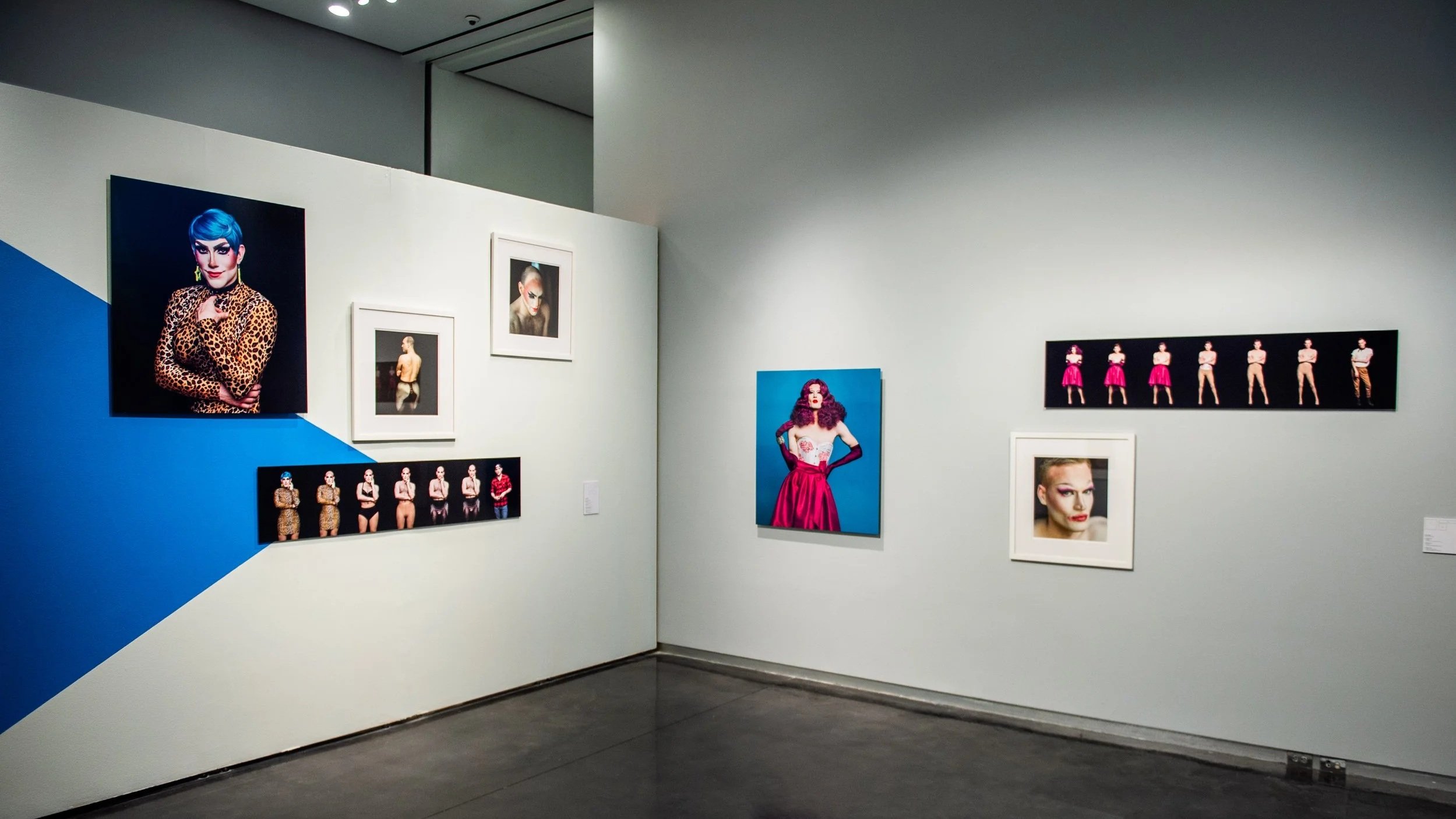 Gallery corner with portrait photographs of individuals in bold makeup and clothing, including a central image of a person in a red dress and a horizontal series showing figure variations by photographer Jerry Siegel.