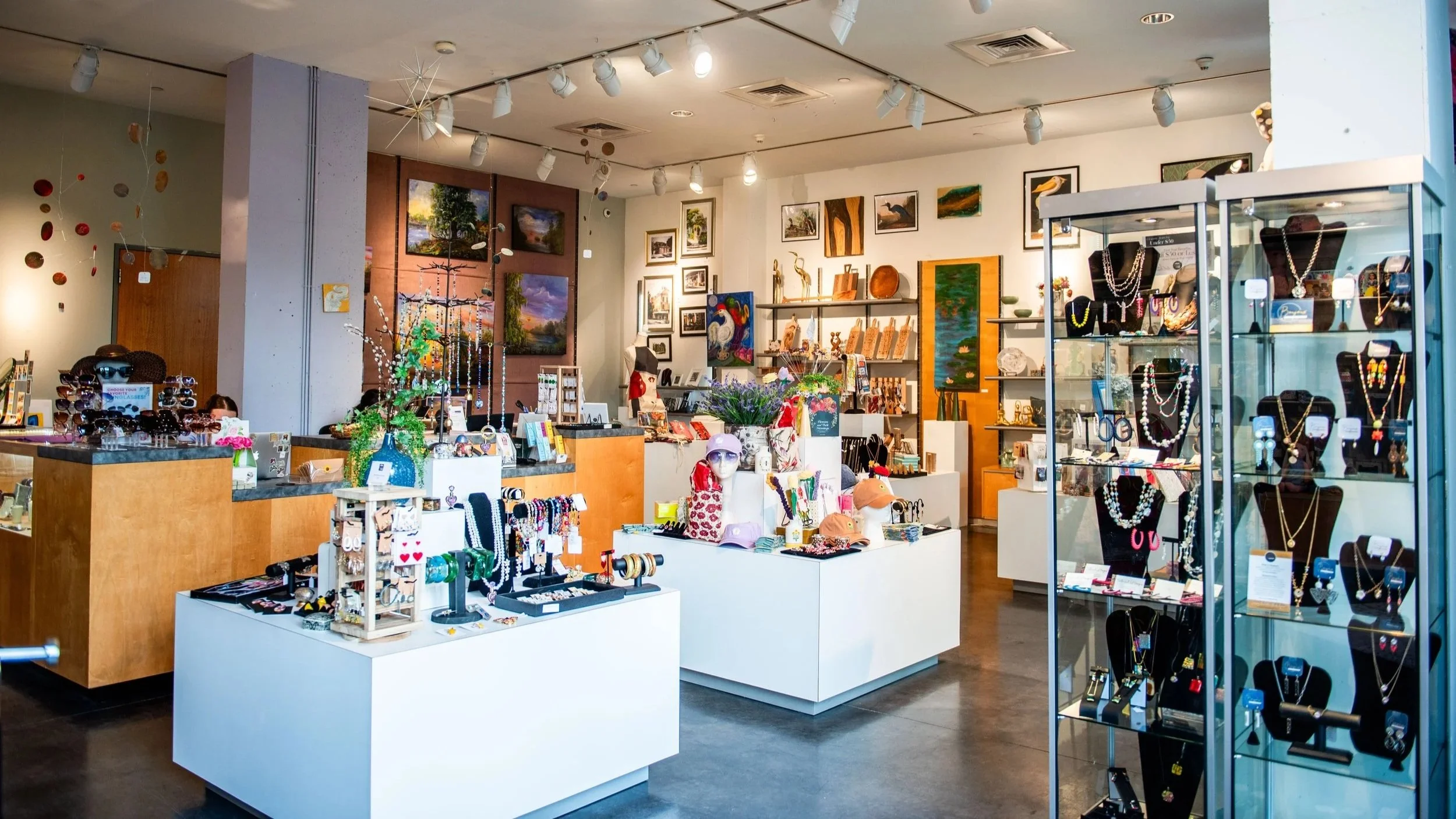 Wide view of a museum gift shop with display tables and glass cases featuring handmade jewelry, accessories, and small artworks, with colorful pieces on the walls and shelves.