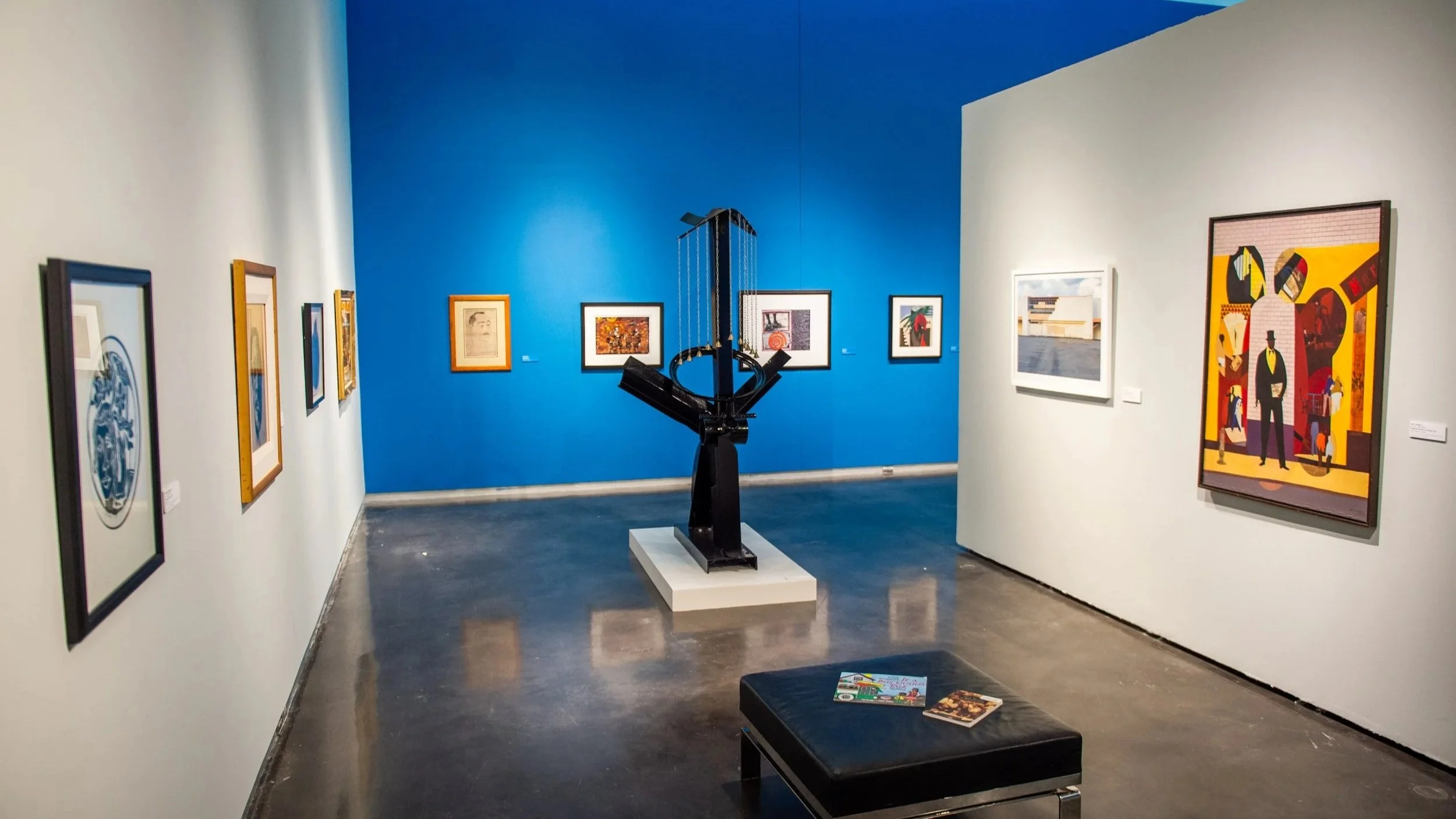 Gallery view with a black sculptural piece on a pedestal centered against a blue wall, surrounded by framed artworks and a bench in the foreground.