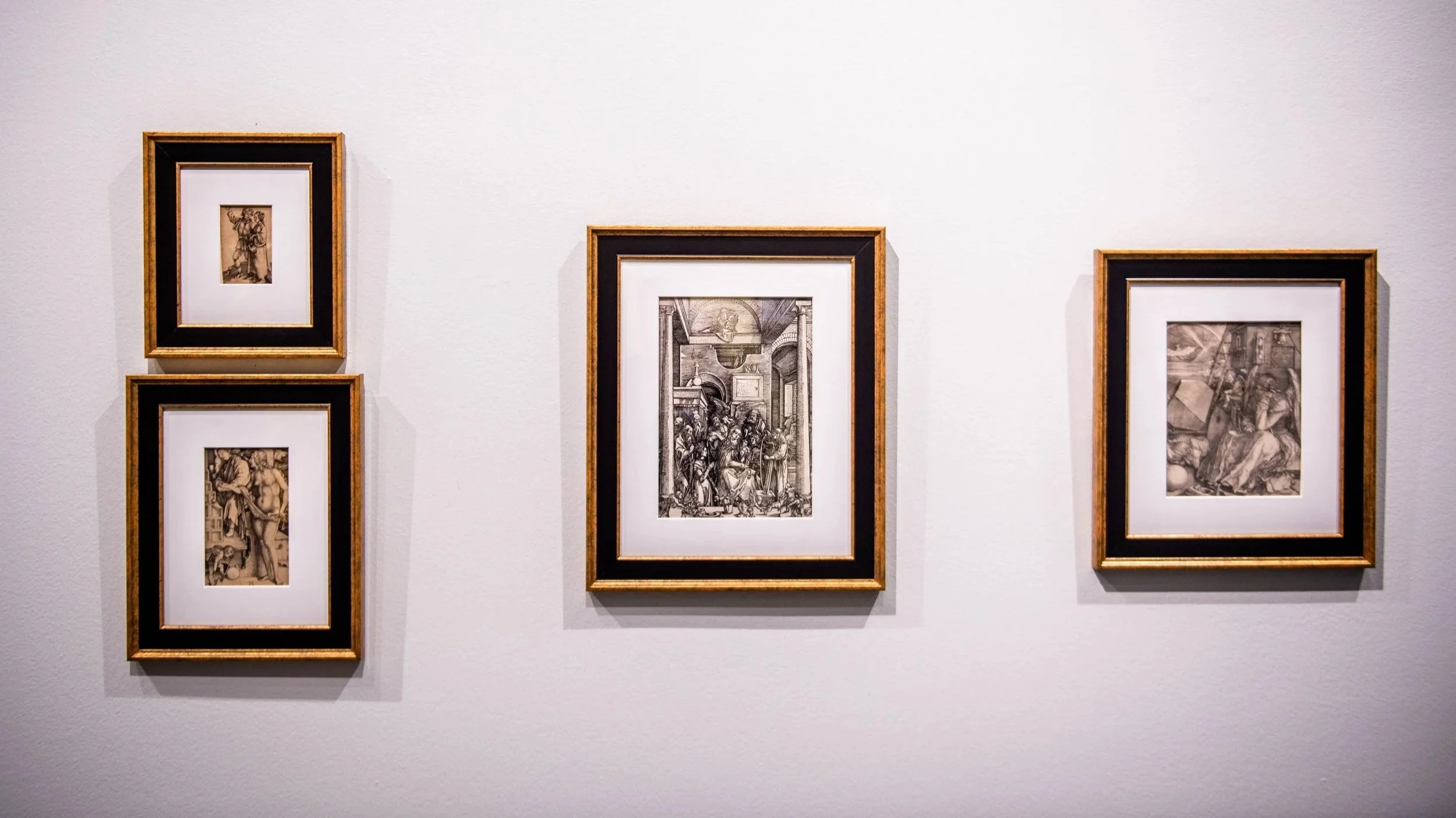 Three framed black-and-white prints hang on a white gallery wall, with two smaller works on the left and a larger central print flanked by another on the right.
