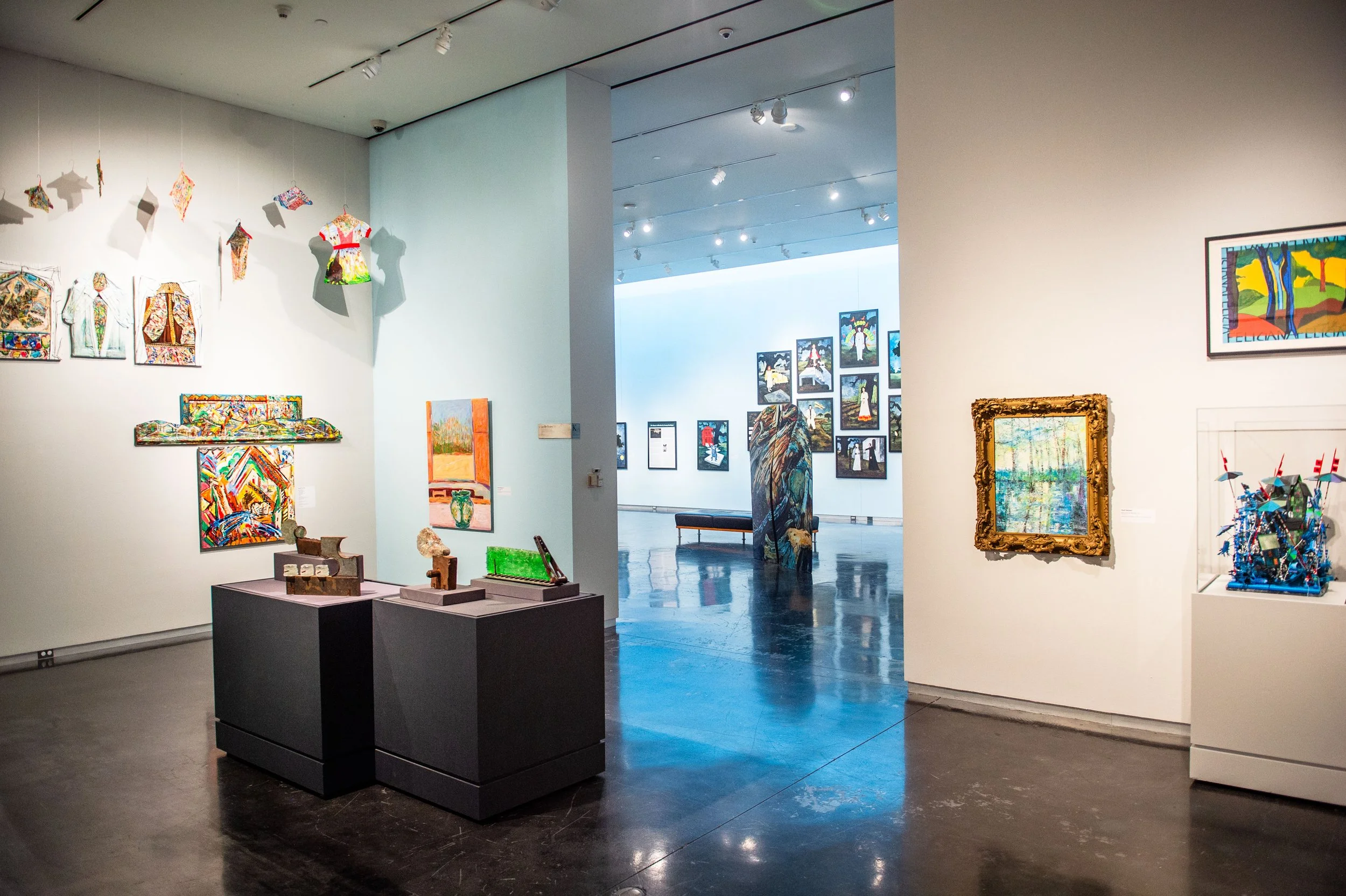Gallery view with colorful mixed-media artworks on walls and small sculptures on display tables, with additional artworks visible in an adjacent gallery through an open doorway.