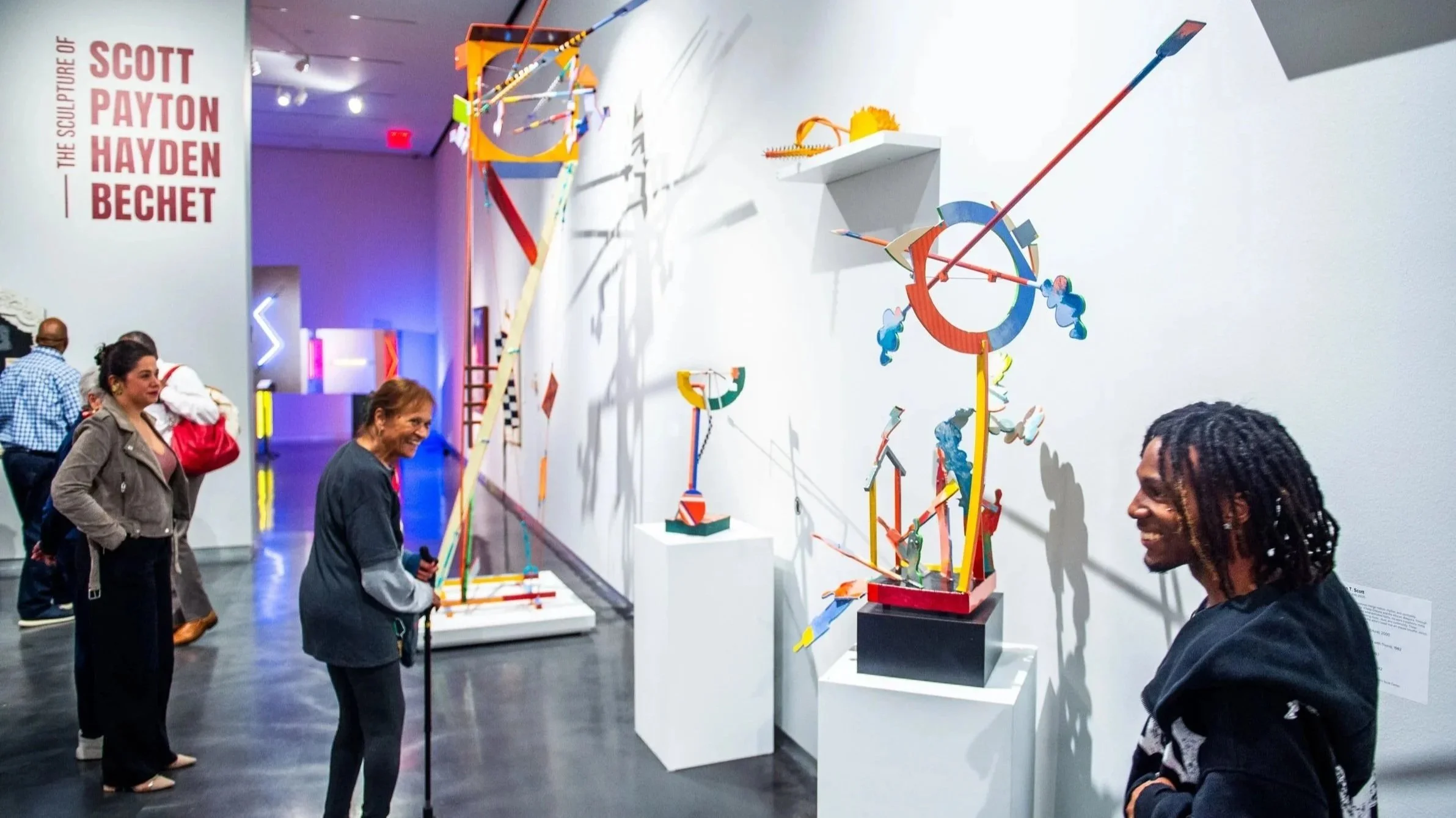 Visitors in a museum gallery smiling and viewing colorful abstract sculptures displayed on pedestals along a wall.