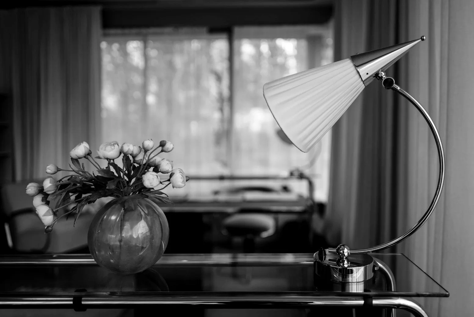 Huis Sonneveld in Rotterdam: interieur met een bloemenvaas met witte bloemen en een moderne bureaulamp op een tafel, met een raam en gordijnen op de achtergrond, in zwart-wit.