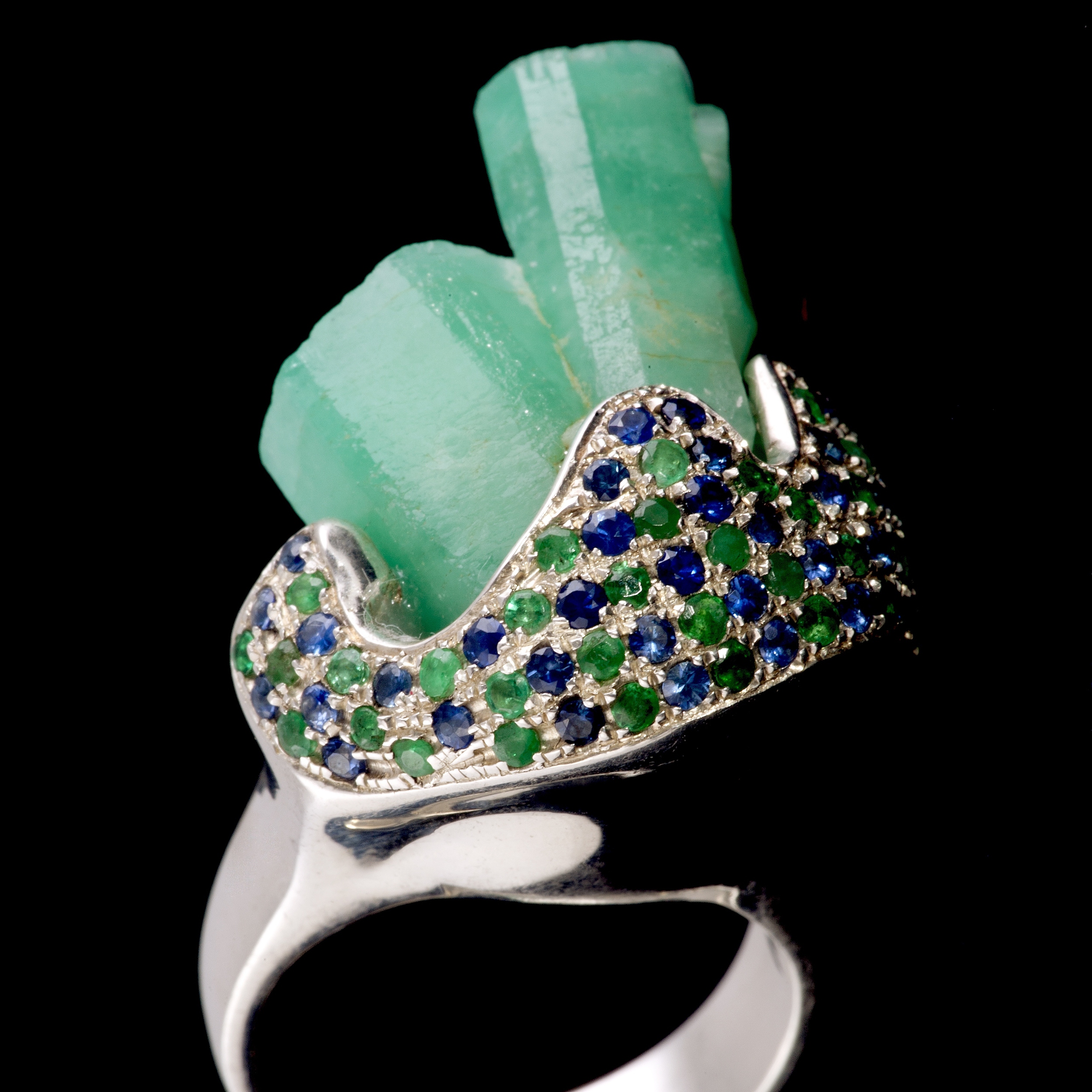 A macle or unique double rough emerald crystals are embraced by sapphire and emerald pavé. Ring is set in 925 silver