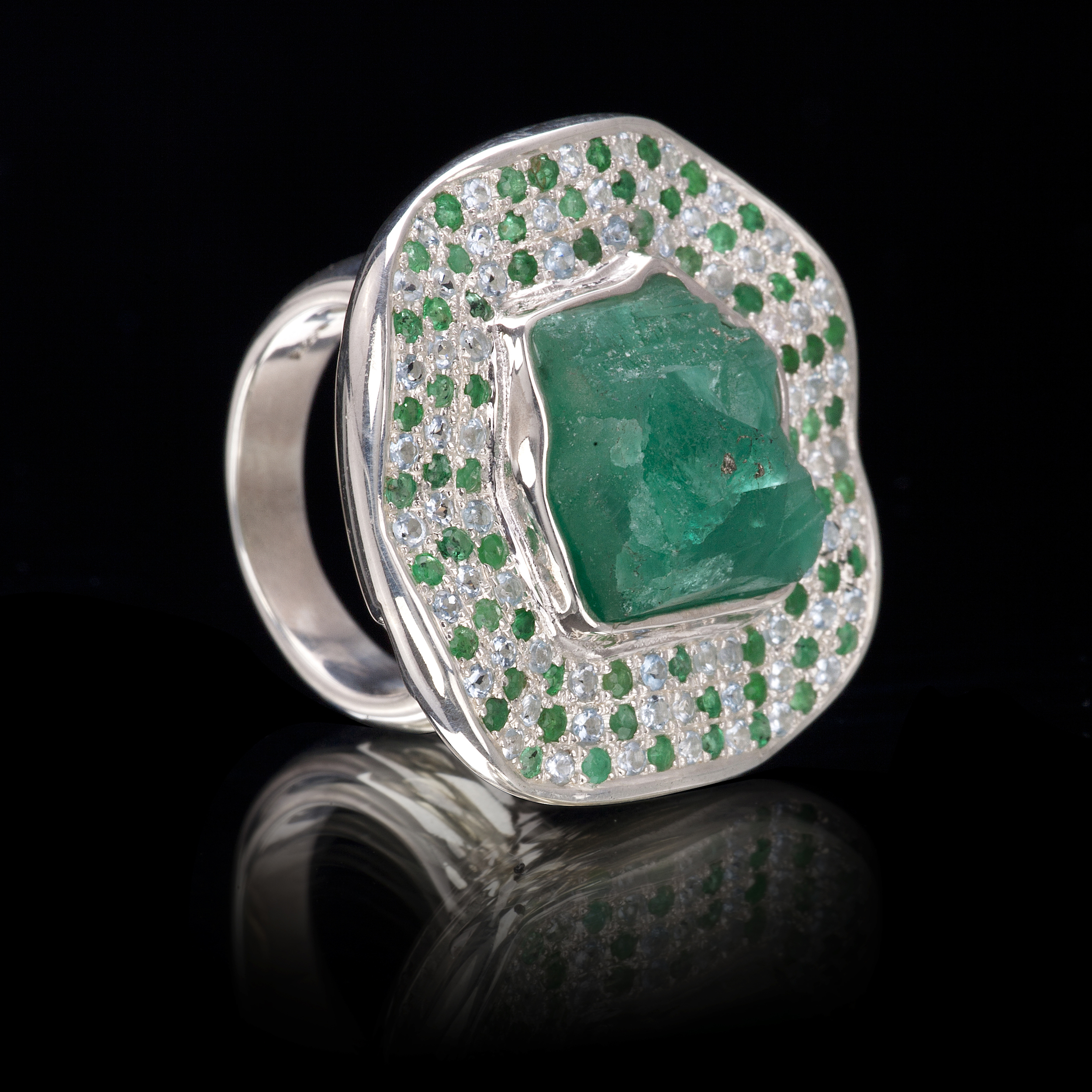 A rough emerald from Colombia is encircled by a wave of aquamarine and emerald pavé. Ring is set in 925 silver
