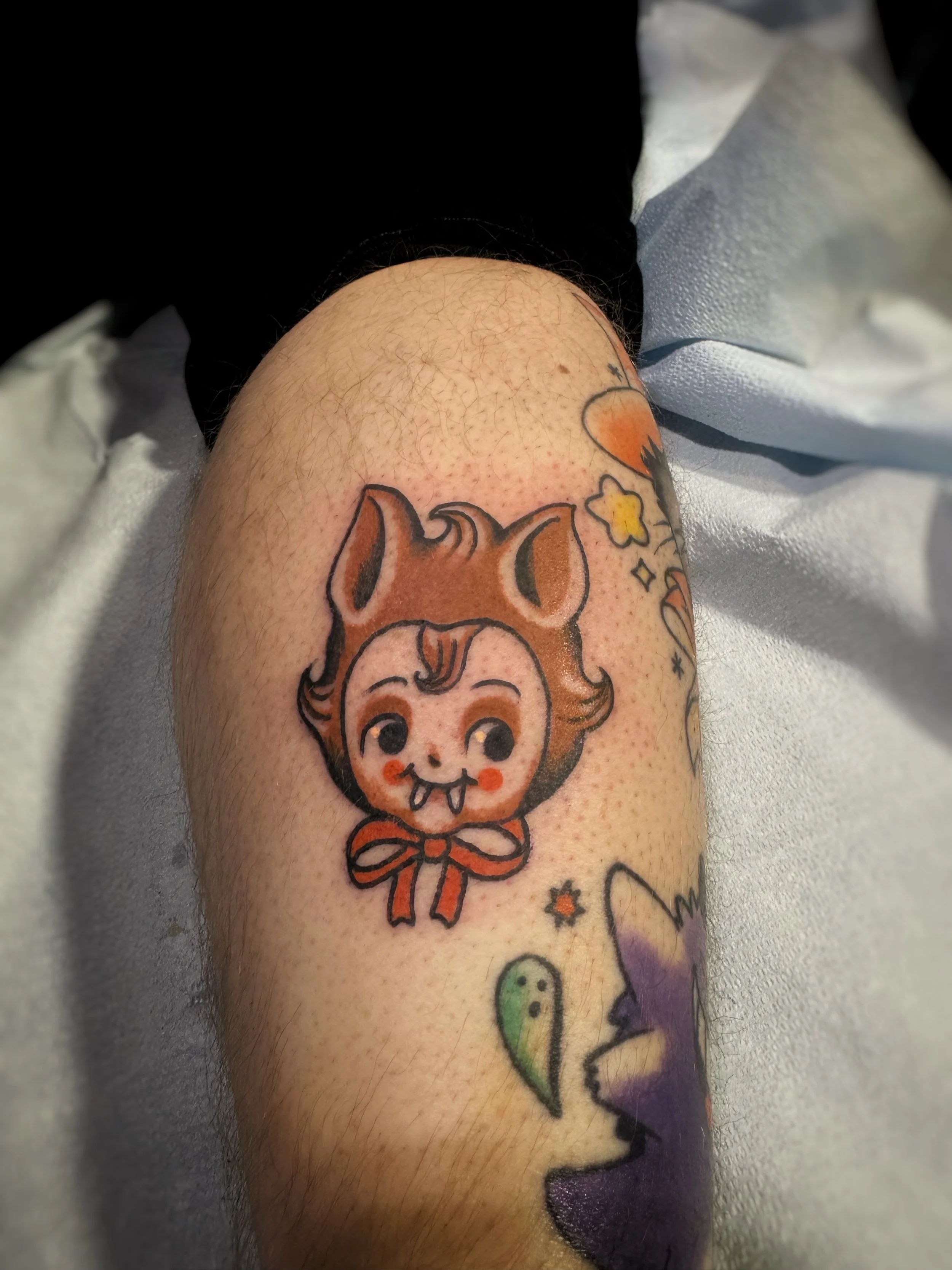 A tattoo of a cartoon character with a skull face, orange hair styled in a vintage look, and a bow around the neck, on a person's arm.