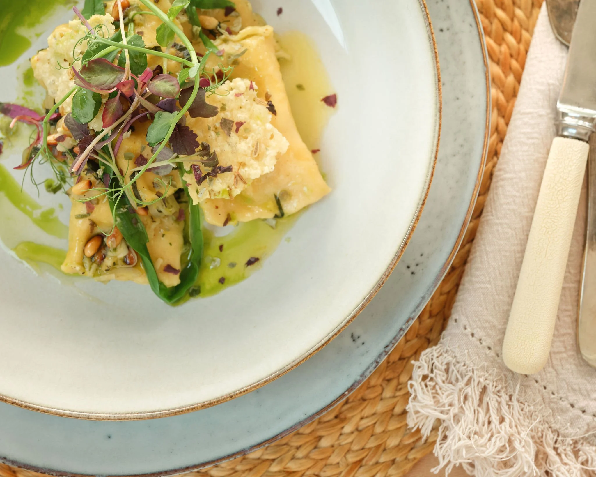 A close-up of a plated dish with pasta, topped with microgreens and garnished with herbs and a green sauce on a white ceramic plate.