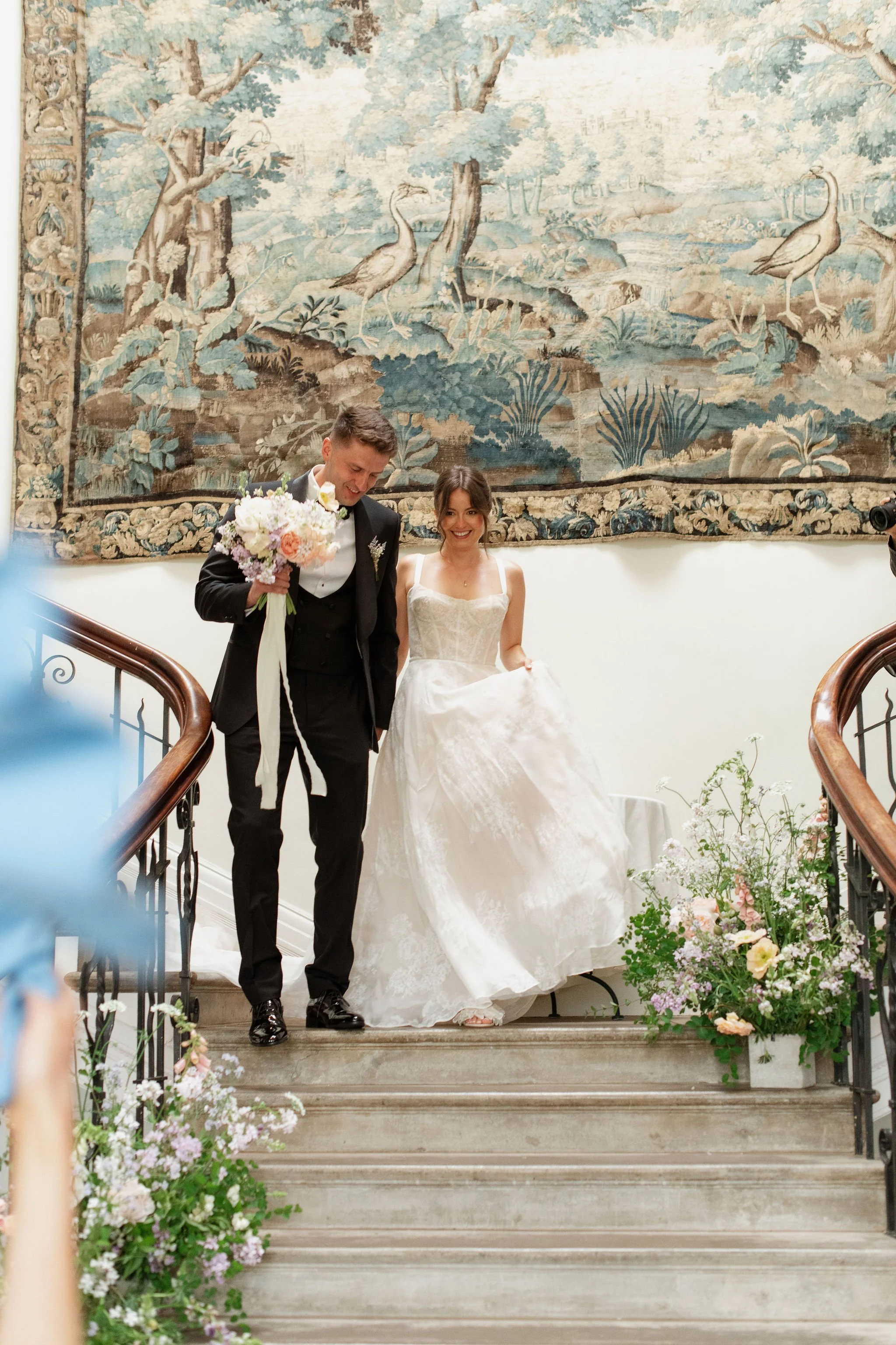 couple with bouquet from The Garden Gate flower company descend a grand light filled staircase at Scorrier House