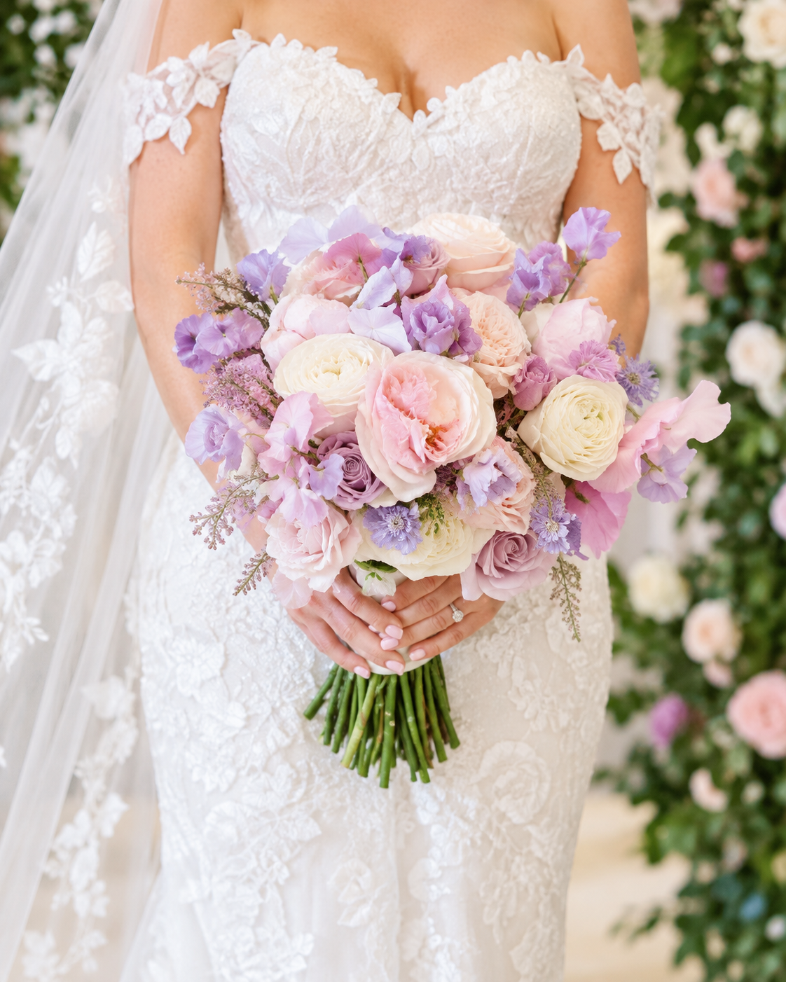 A bride in a white lace wedding dress holding a bouquet of pink, cream, and lavender flowers.