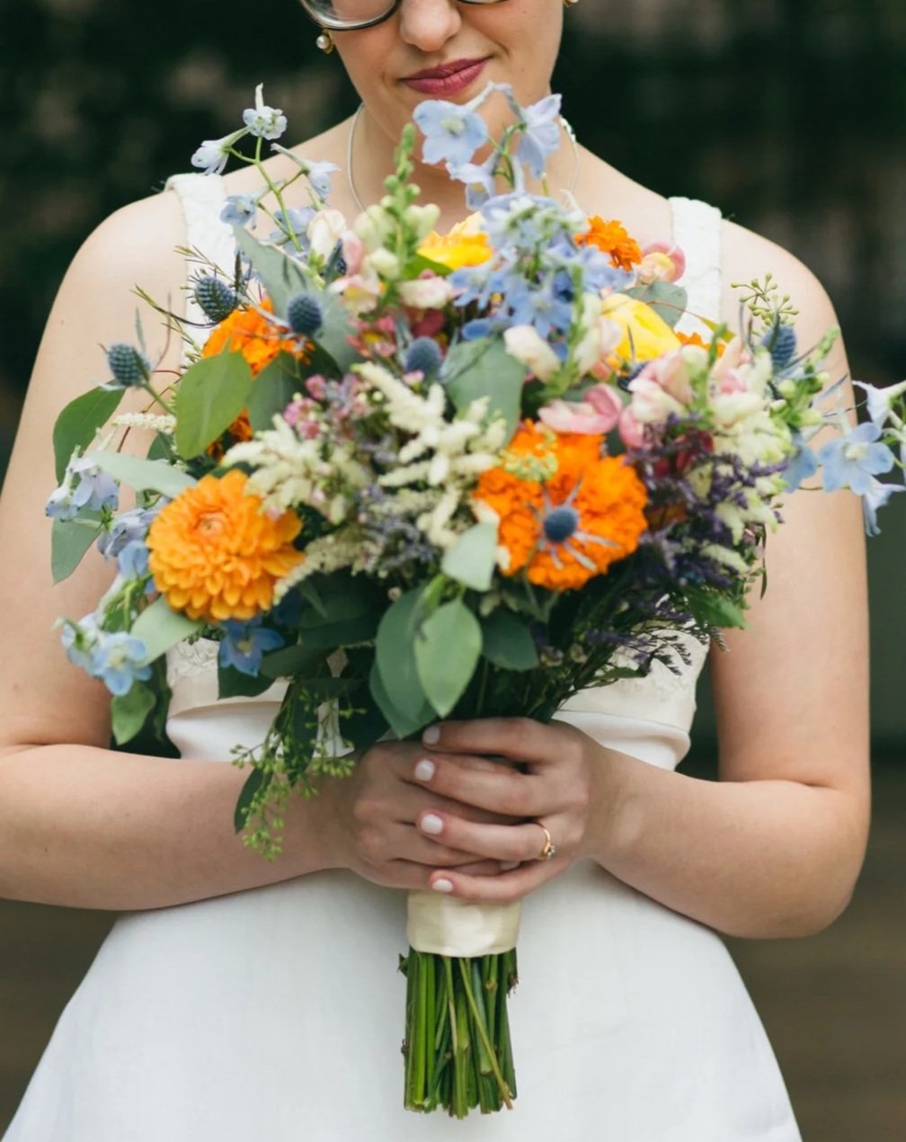 A woman in a white sleeveless dress holding a colorful bouquet of mixed flowers, including marigolds, blue thistles, and various greenery.
