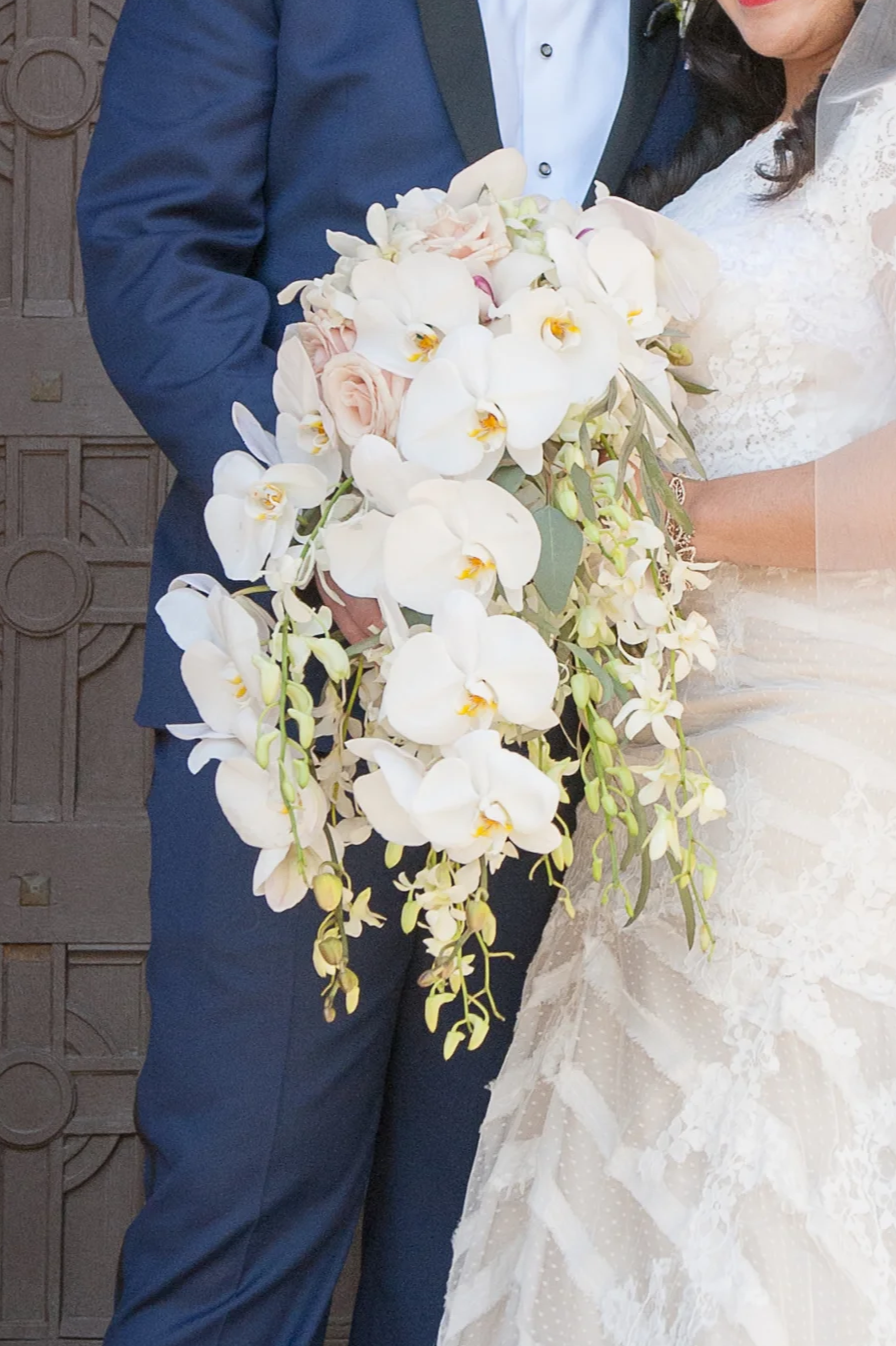 Close-up of a bride and groom holding a cascading wedding bouquet of white orchids, pale pink roses, and greenery in front of a textured wooden door.