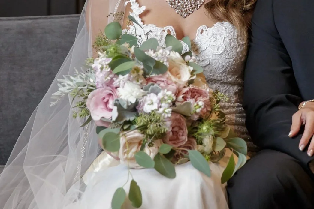 Close-up of a bride holding a bouquet of pink, white, and green flowers, sitting next to someone in a black suit.