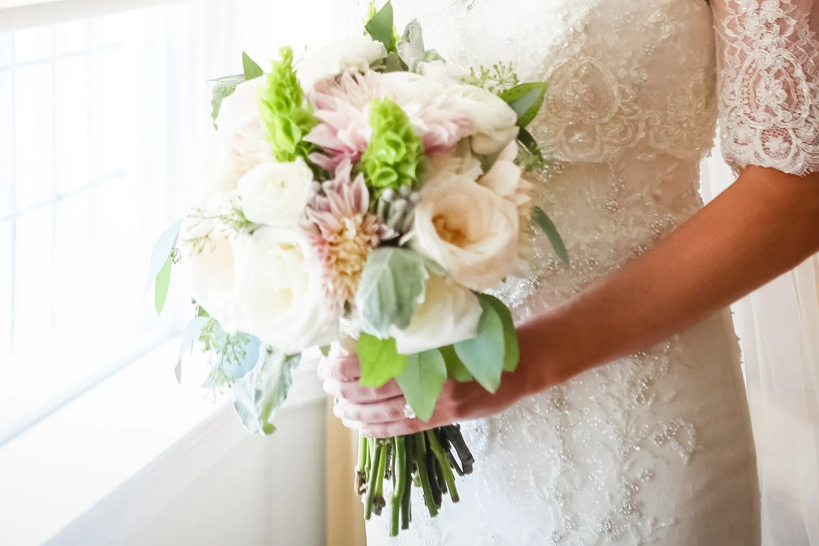 A bride holding a bouquet of white and light pink flowers with green leaves, wearing a lace wedding dress, standing near a window.