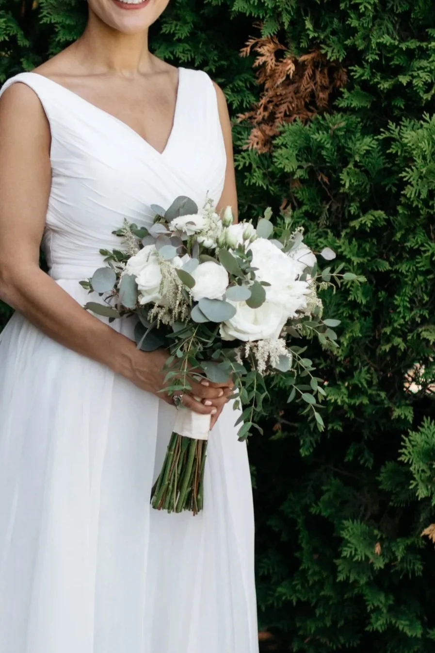 A woman wearing a white wedding dress holding a bouquet of white flowers and greenery, standing in front of green foliage.