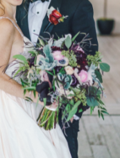 Bride and groom holding a large bouquet of purple, white, and green flowers.