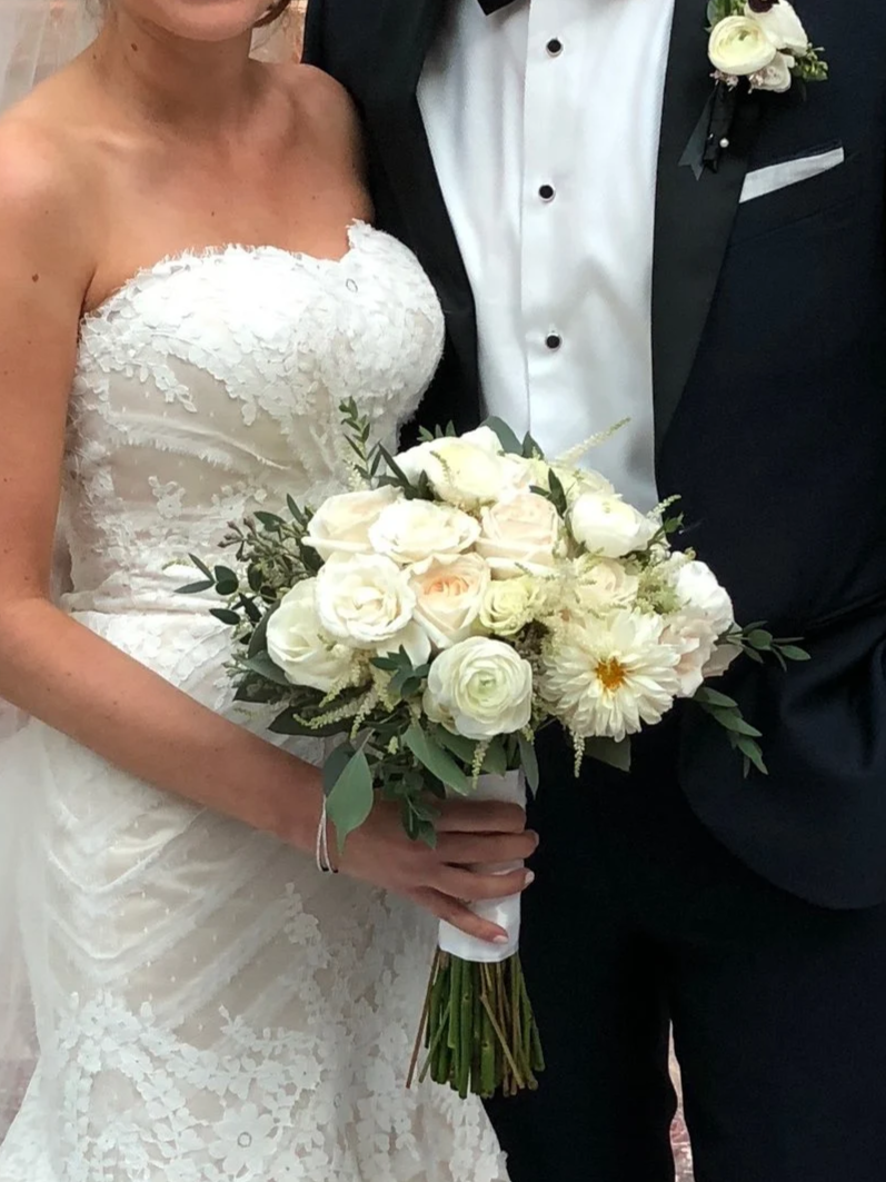 Close-up of a bride and groom holding a bouquet of white roses, peonies, and greenery, dressed in wedding attire.