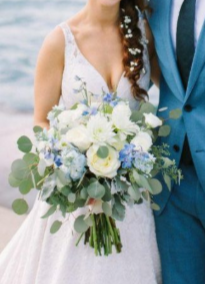 Bride in a white wedding dress holding a bouquet of white and blue flowers, next to a person in a blue suit.