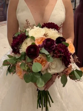 A person in a white dress holding a vibrant bouquet of flowers with white, dark red, orange, and purple blooms, along with green foliage.