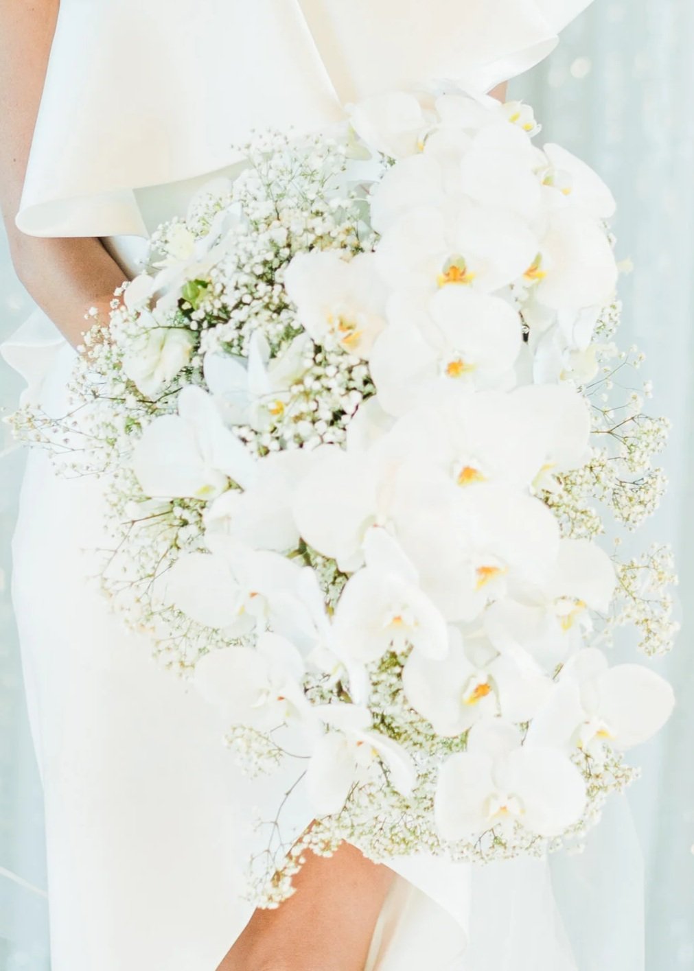 Bride holding a cascading white orchid and baby's breath bouquet in a white wedding dress.