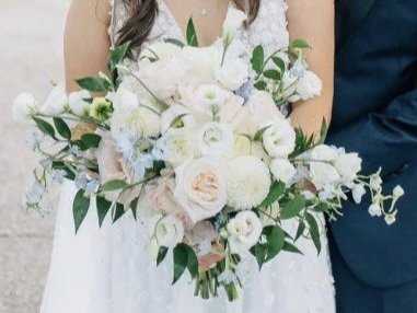 A bride holding a large bouquet of white and light pink flowers with green leaves, standing next to a person dressed in dark blue.