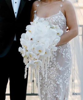 Bride in a white floral lace wedding dress holding a cascading bouquet of white flowers, with groom dressed in a tuxedo beside her.