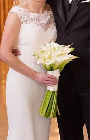Bride in a white wedding dress holding a bouquet of white calla lilies, standing next to a groom in a dark suit.