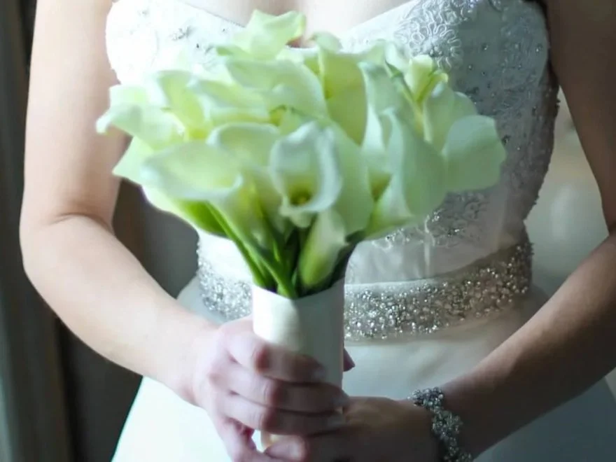 A bride holding a bouquet of white calla lilies, dressed in a white wedding gown with embellishments.