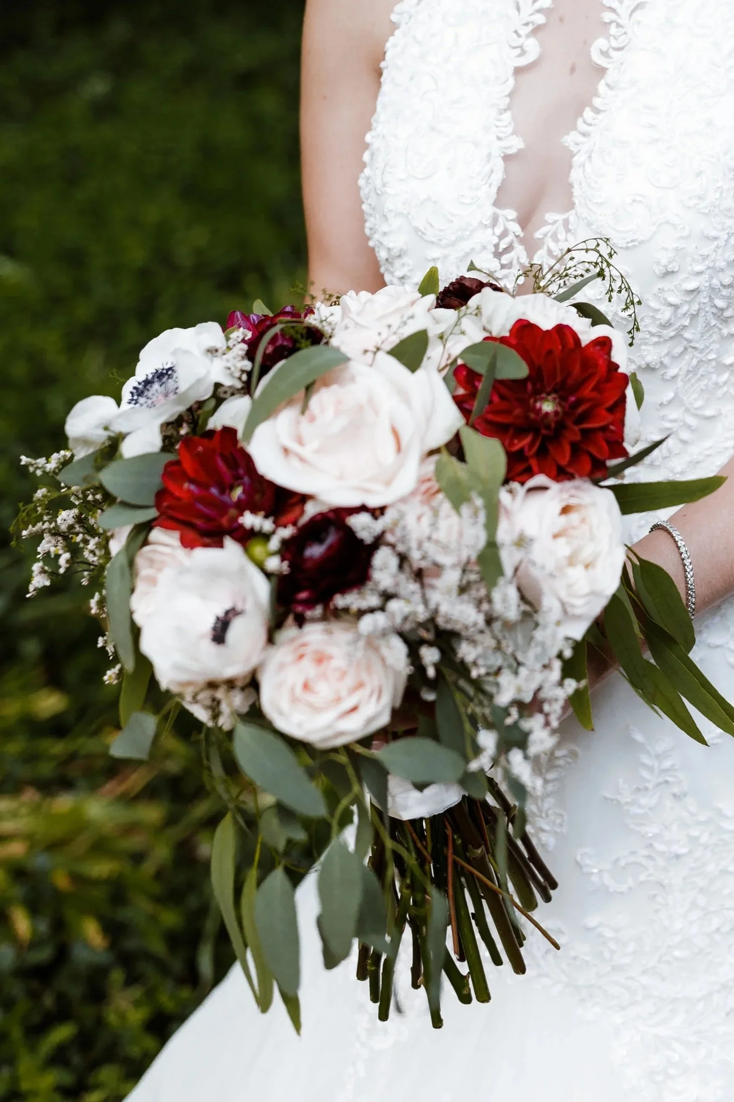 Close-up of a bride holding a bouquet of white, red, and dark purple flowers with green leaves. The bride is wearing a white dress with intricate lace and embroidery details.
