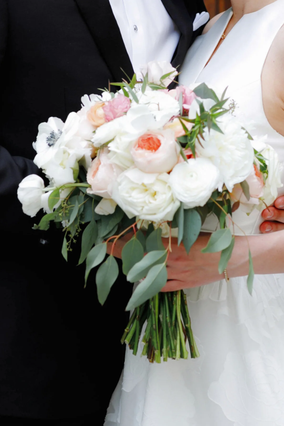 Close-up of a bride and groom holding a bouquet of white, pink, and peach flowers with green foliage at their wedding.