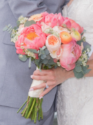 A person holding a bouquet of pink, peach, and white flowers with greenery, dressed in a gray suit.