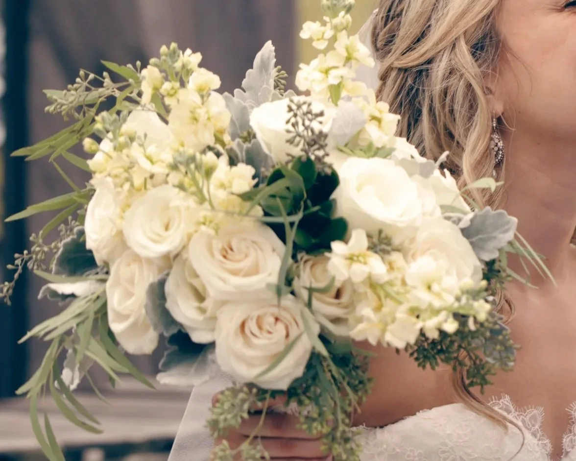 A bride holding a bouquet of white and cream roses, with green and gray foliage, next to her face and shoulder.