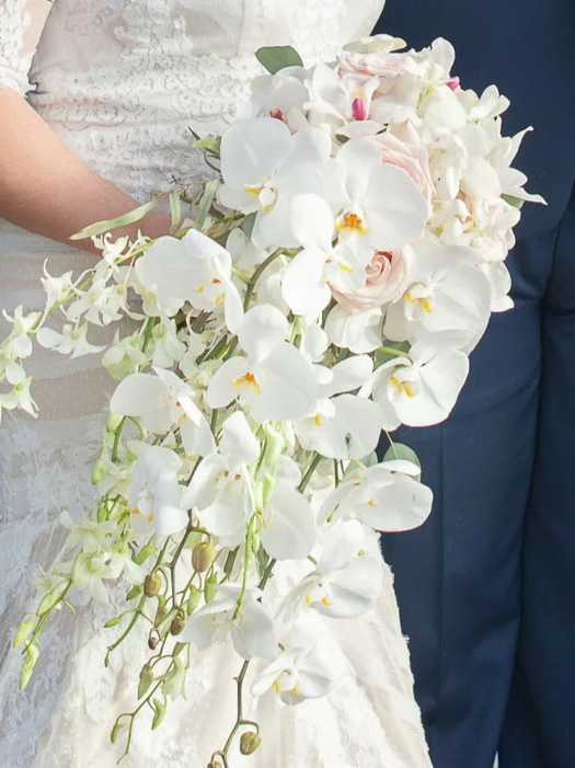 Bride holding a cascading bouquet of white orchids and pale pink roses, with a lace wedding dress and someone in a navy blue suit in the background.