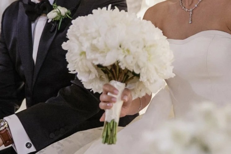 Close-up of a bride holding a bouquet of white flowers, with a groom in a tuxedo beside her during a wedding.