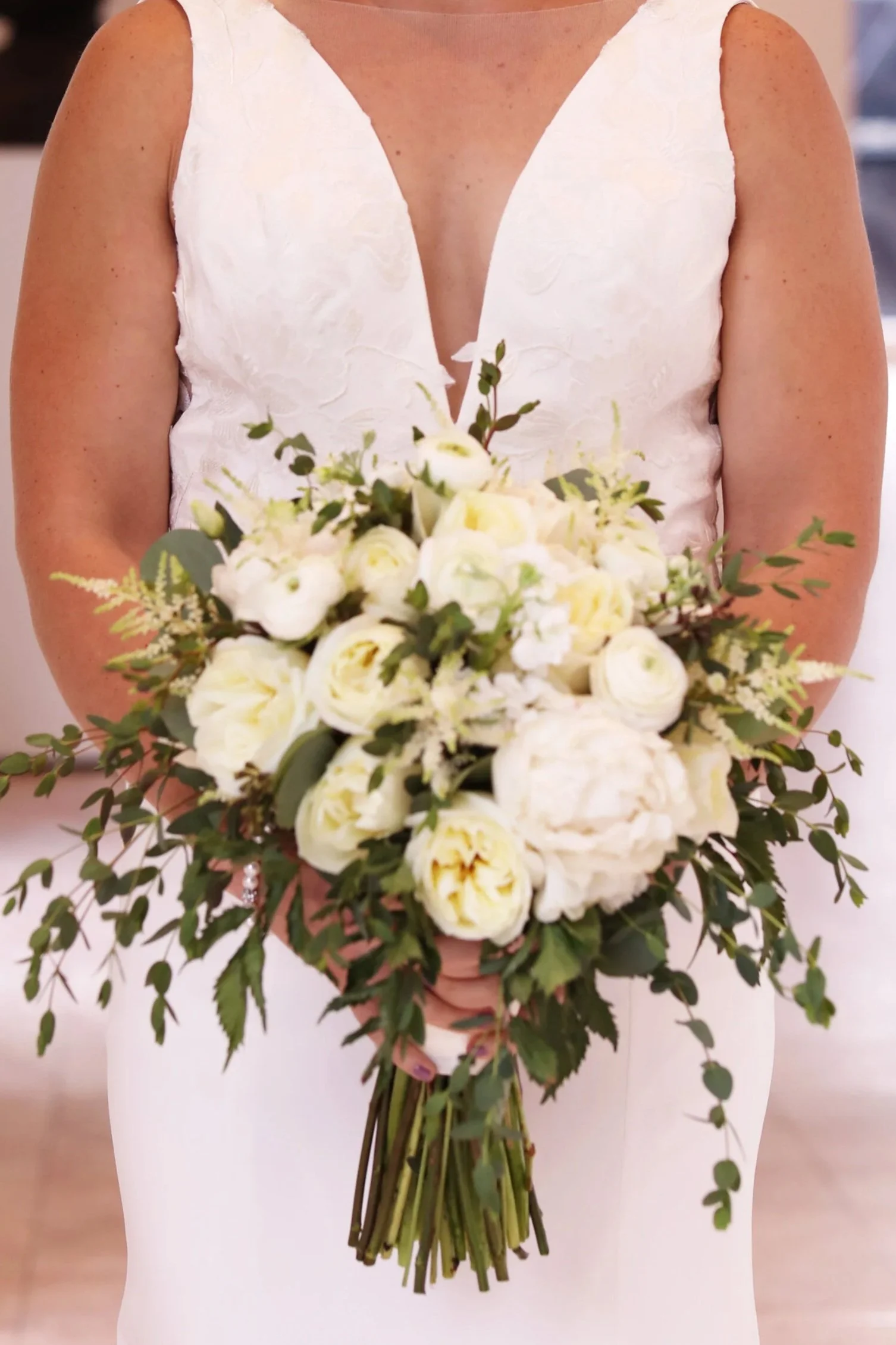 A woman in a white dress holds a large bouquet of white flowers and greenery, standing indoors.