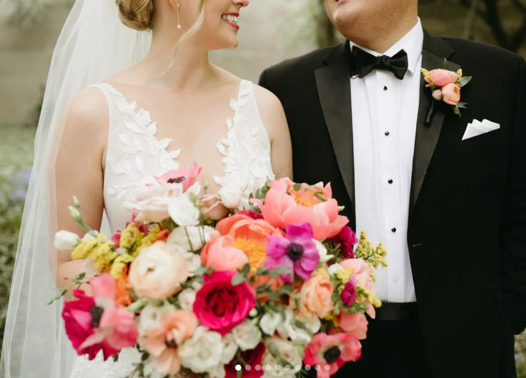 A bride and groom on their wedding day, with the bride holding a vibrant bouquet of pink, peach, and white flowers. The bride wears a white lace wedding dress, and the groom is in a black tuxedo with a bow tie and boutonniere.