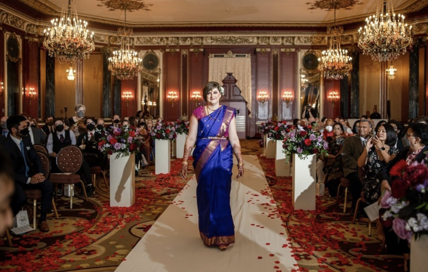 A woman in a blue and pink saree walking down an aisle at a wedding ceremony in an ornate, chandelier-lit hall decorated with flowers and red rose petals.