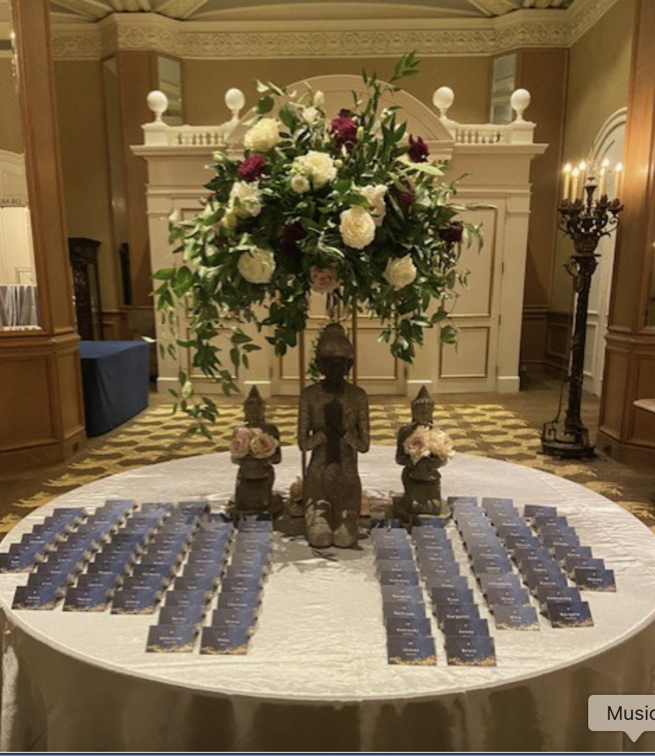 A round table with a large floral arrangement featuring white and pink flowers, decorative statues including a large central Buddha figure, and multiple cards or place cards organized around the table, located in a richly decorated room with ornate m
