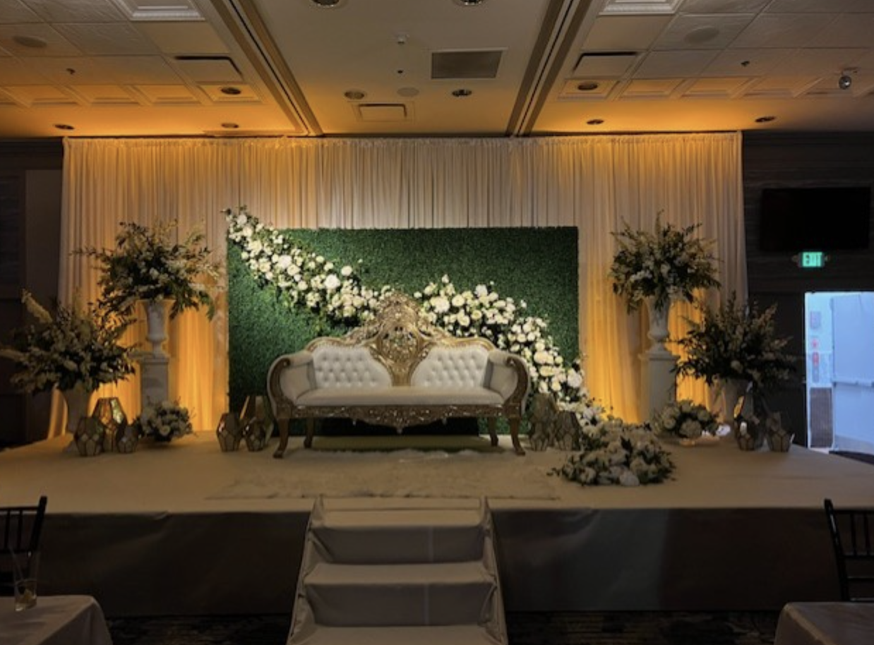 Elegant decorated stage with a white vintage sofa, floral arrangements, and a green backdrop with white flowers for a wedding or special event.