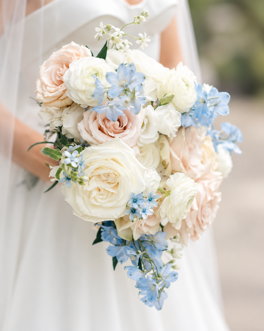 A wedding bouquet featuring white, blush pink, and blue flowers held by a bride in a white dress.