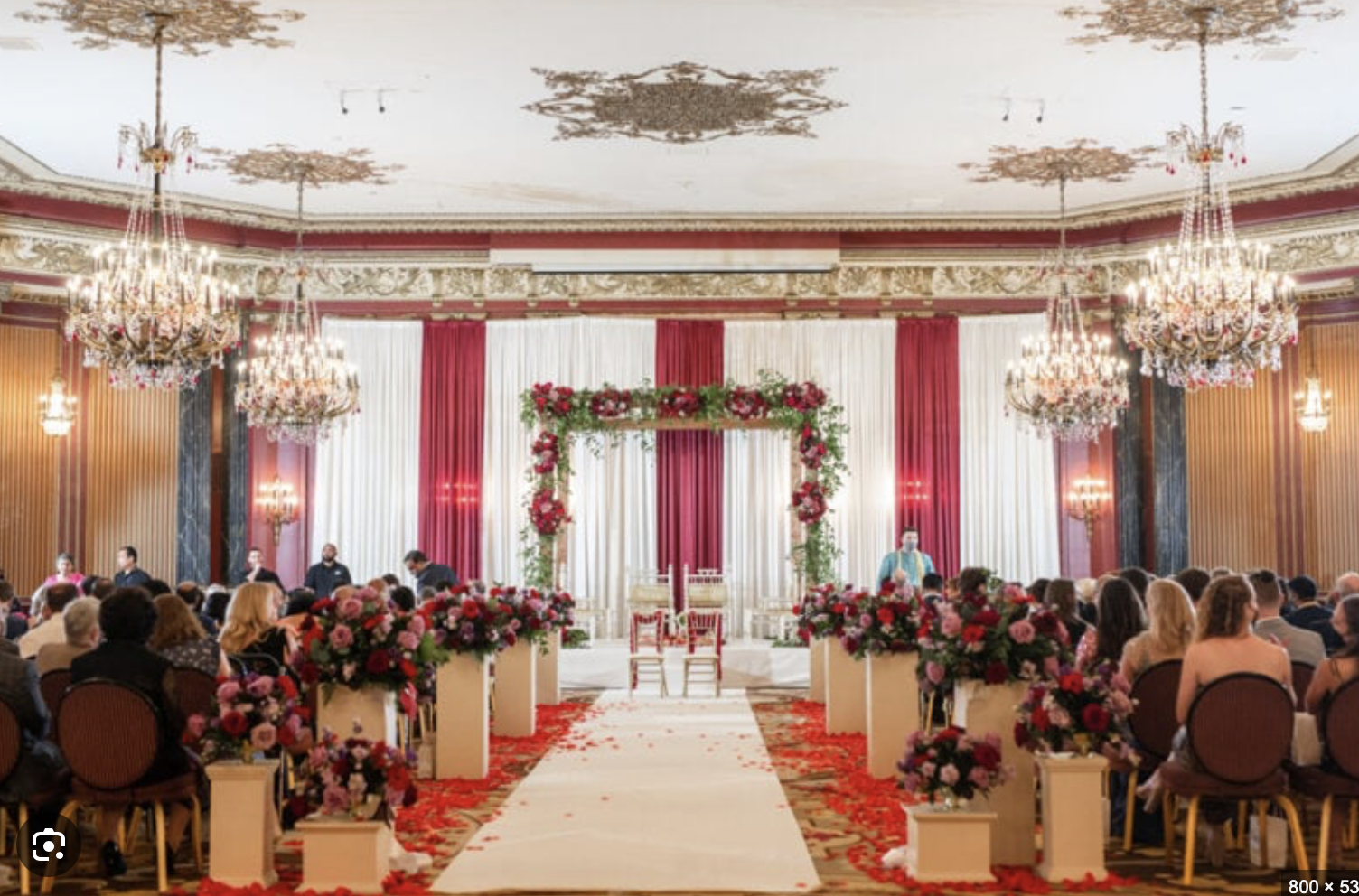 Elegant wedding ceremony setup with floral arch, white chairs, and red and pink flower arrangements in a grand hall with chandeliers and ornate ceiling details.
