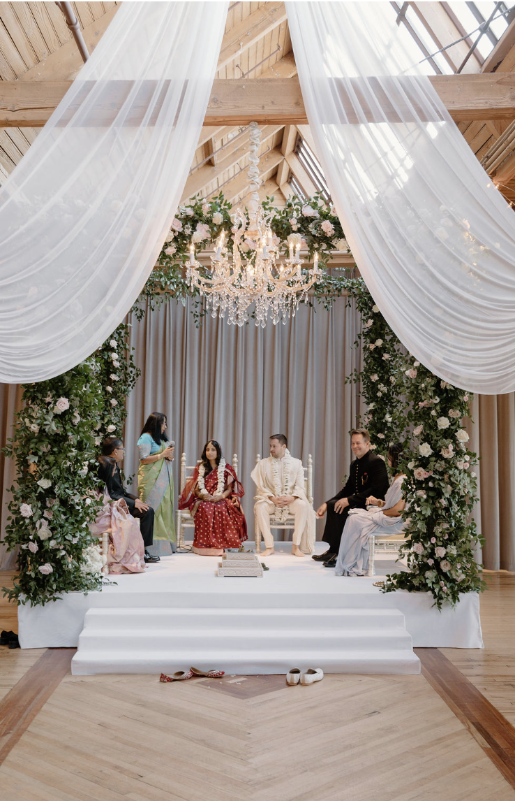 A traditional Indian wedding ceremony with four people sitting on a stage decorated with floral arrangements and drapery, under a chandelier in a wooden venue.
