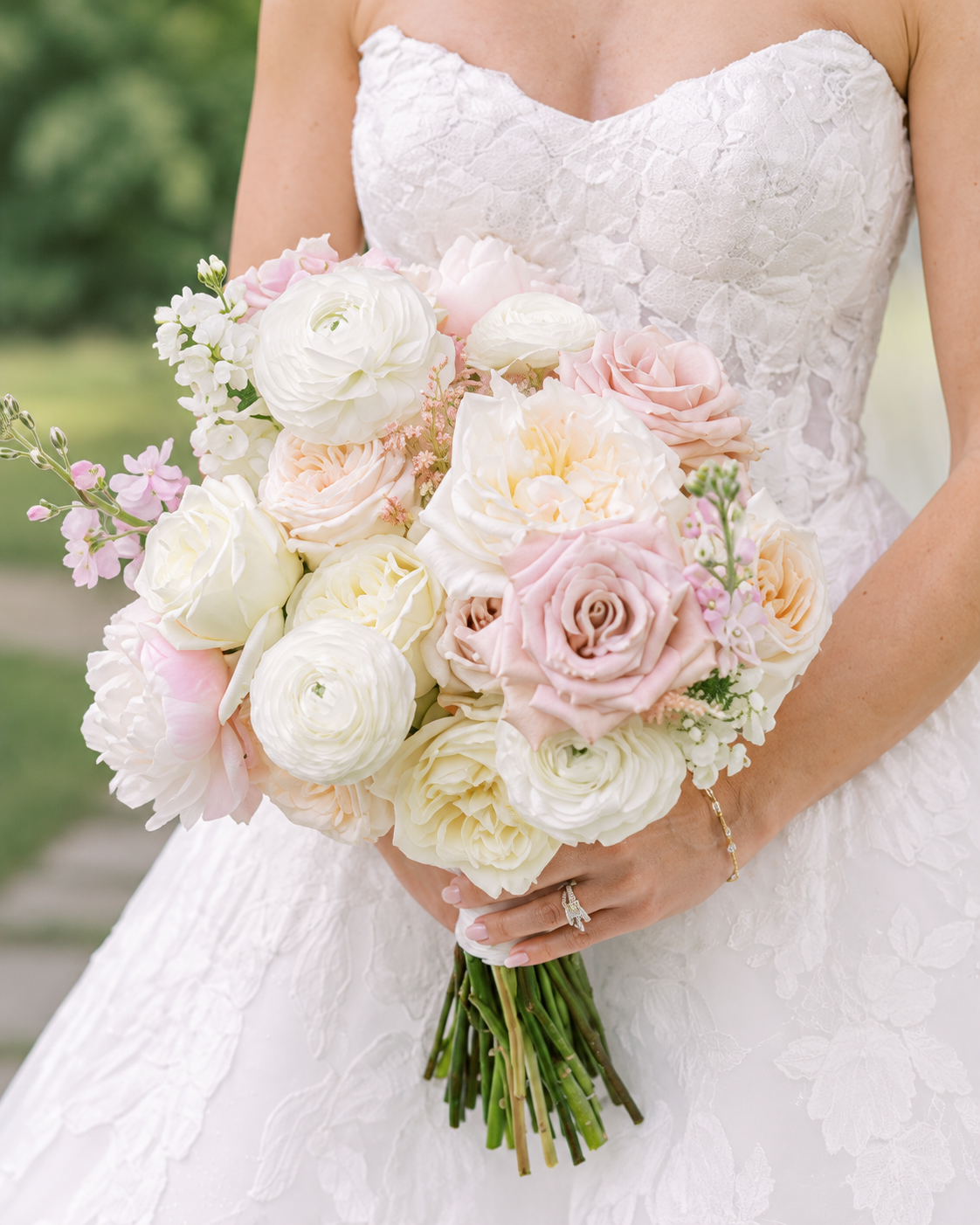 A bride holding a bouquet of white, cream, and pink roses and peonies in a garden setting.