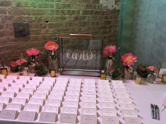 Table with small favor boxes, pink flowers, and a sign that says 'Cloe' in a glass frame, set against a brick wall.