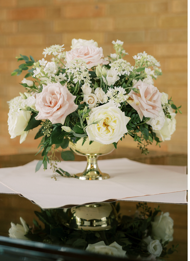 A floral arrangement with pink and white roses and other white flowers in a gold vase on a table.