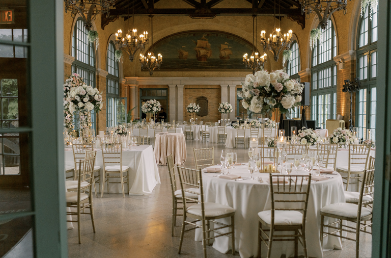 Elegant event space with round tables covered in white tablecloths, decorated with floral centerpieces and lit candles, set for a formal gathering or wedding reception.