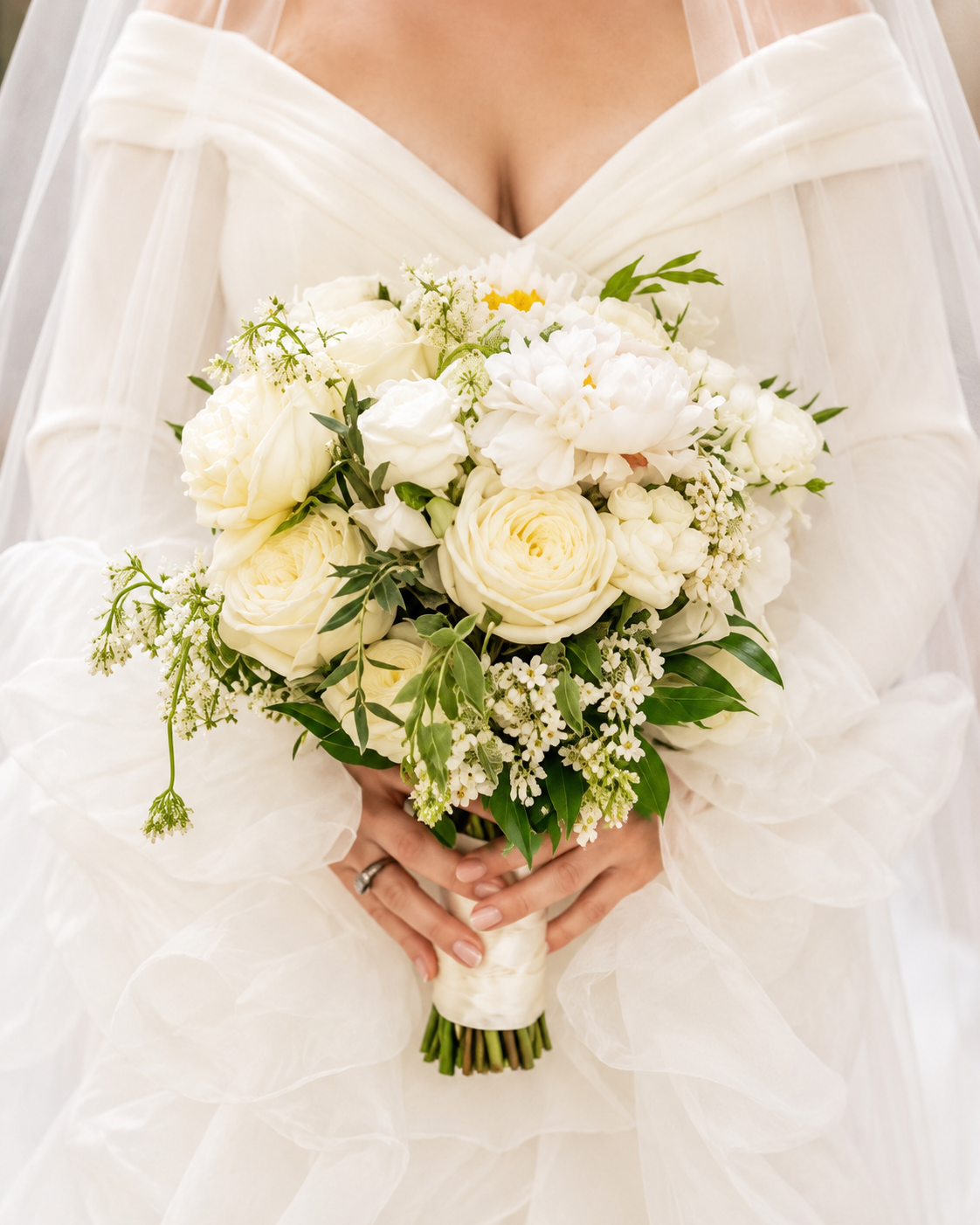 Bride holding a bouquet of white roses, peonies, and greenery in front of her white wedding dress.
