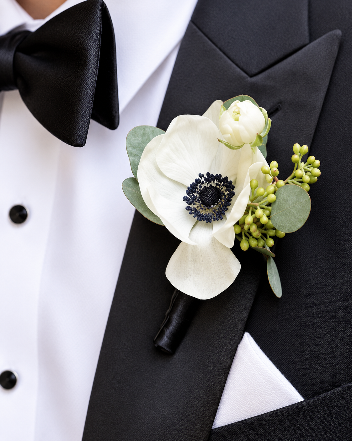 Close-up of a tuxedo with a white shirt, black bow tie, and a boutonniere featuring white anemone flowers, green leaves, and berries.