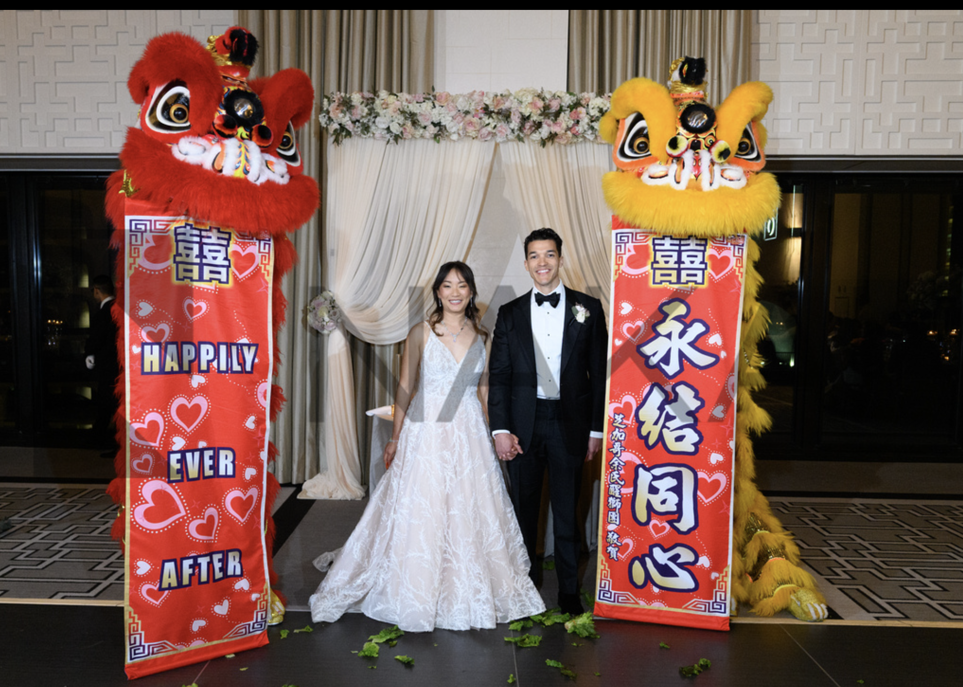 A bride and groom holding hands, standing between two large red and yellow Chinese lion dance costumes at a wedding celebration. There are decorative banners with hearts and Chinese characters behind them. The bride is in a white wedding gown, and th