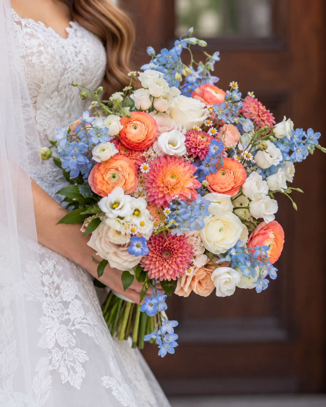 A bride holding a colorful bouquet of various flowers including orange dahlias, white roses, blue delphiniums, and small daisies, partially visible with lace wedding dress and veil.